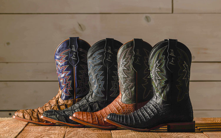 Four boots lined up in a row with their toes to the left and heels to the right with a spotlight on them and placed in front of a wooden backdrop.