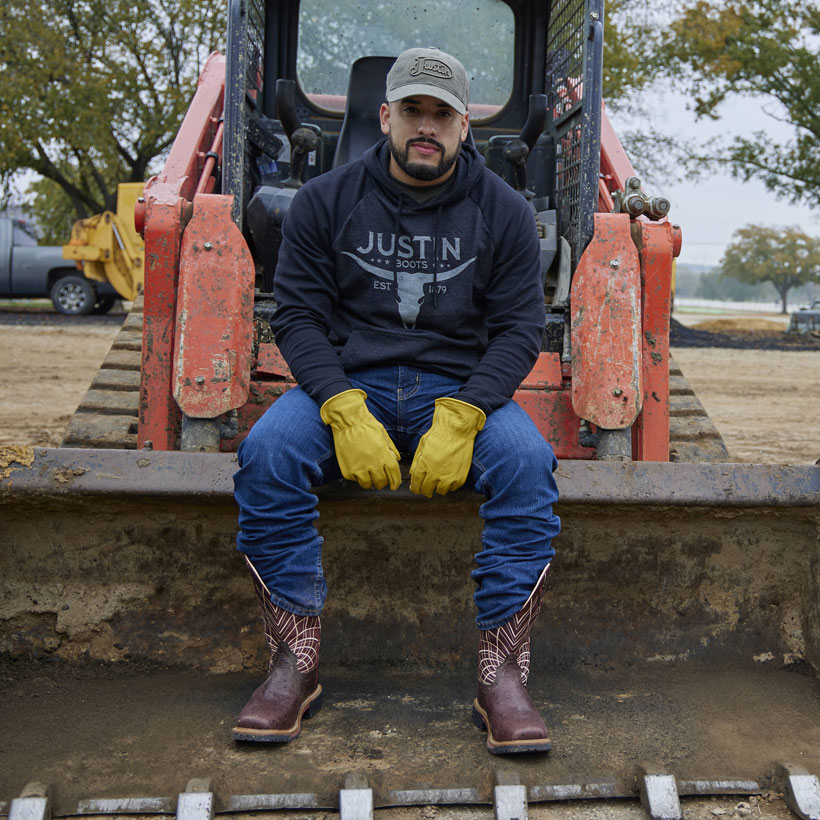 A man wearing Derrickman 12" Waterproof work western boots in brown and Performance Fleece Hoodie in grey sitting on a tractor bucket.