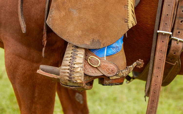 A cowboy’s boot is seen riding a horse with his foot in the stirrup with a spur and a blue top.