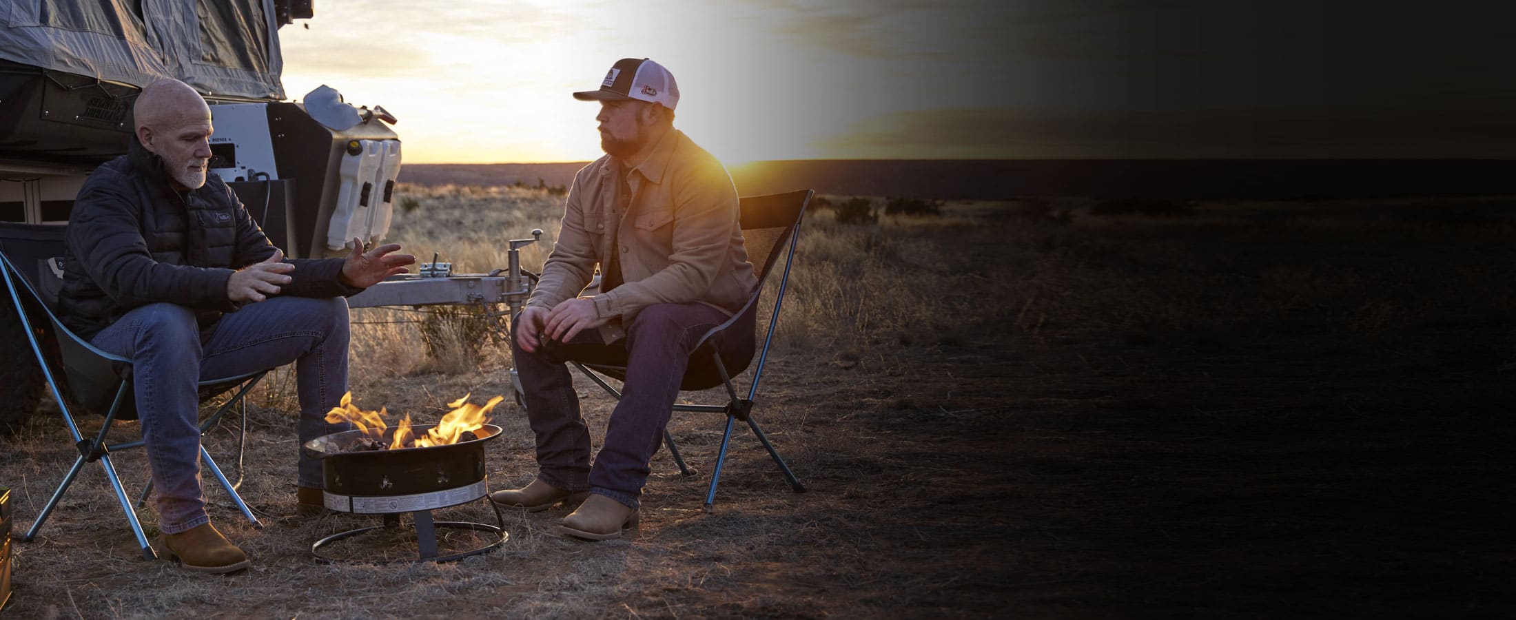 Two pairs of men’s Big Bucks 11” Western boots in brown worn by two men, posing next to a campfire. 