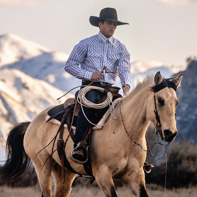 A cowboy wearing the Justin Big News 11-inch Western Boots in Walnut (brown) while riding a horse. Shop Big News.