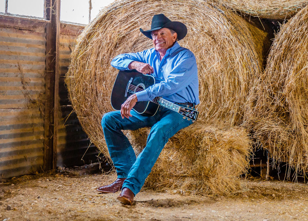 A man wearing Justin George Strait western boots, a cowboy hat and holding a guitar while sitting on a hay bale in a barn.