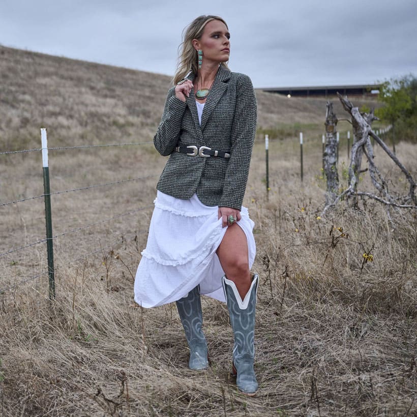 Women’s Verlie 17” women’s western boot in blue grey worn by a blonde woman also wearing a blazer and a white dress, standing in a field.