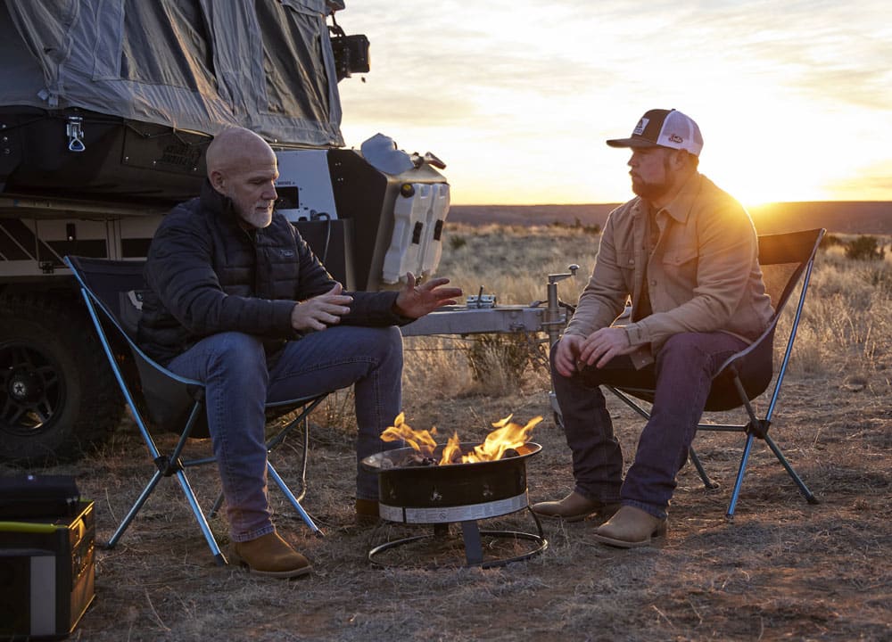 Two pairs of men’s Big Bucks 11” Western boots in brown worn by two men, posing next to a campfire. 