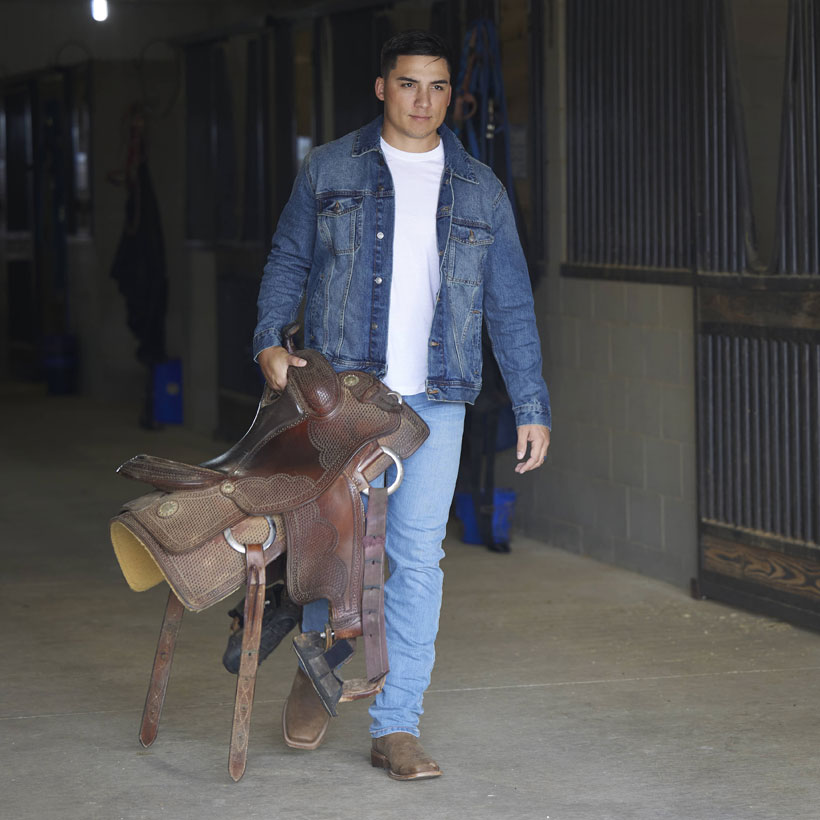 A man wearing Jackpot 11" Western boot in walnut brown carrying a saddle in a barn.