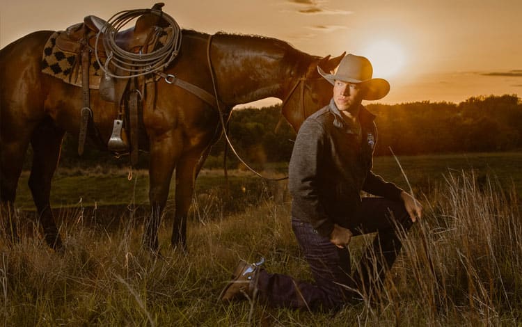 cody con sombrero de vaquero se arrodilla en el campo frente a un caballo