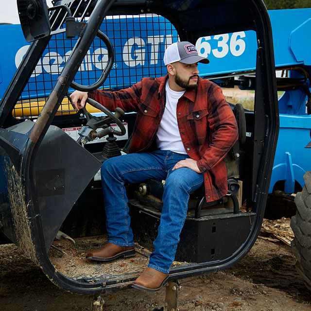 Man seated in the cab of a blue industrial tractor wearing Justin Handler 11-inch Composite Toe Work Boots in Brown. Shop Handler.