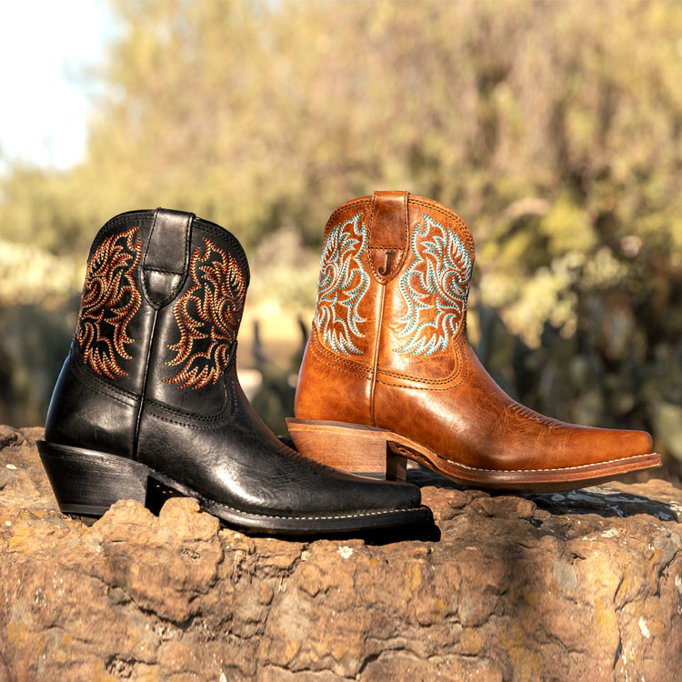 Justin Isabella 7-inch Booties in black and Justin Isabella 7-inch Booties in brown sitting on a rock wall. Shop Isabella.