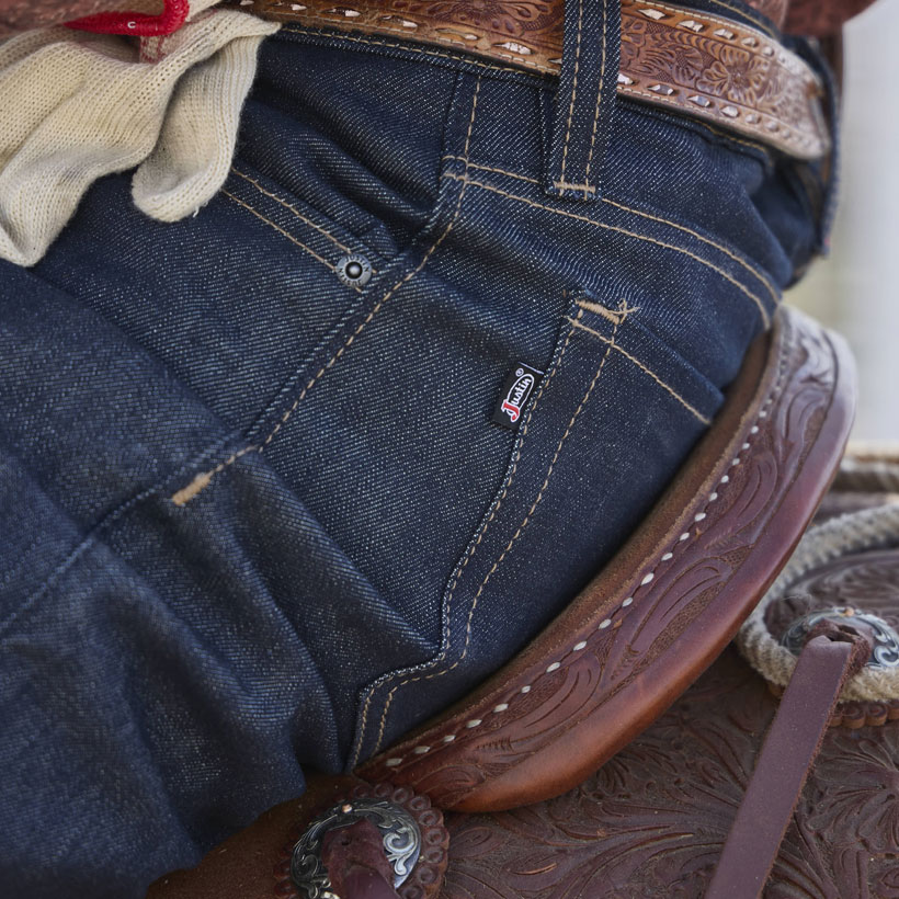 Vaqueros clásicos de pierna recta para hombre en azul oscuro sin lavar, usados ​​por un vaquero sentado en una silla de montar con un guante en el cinturón.