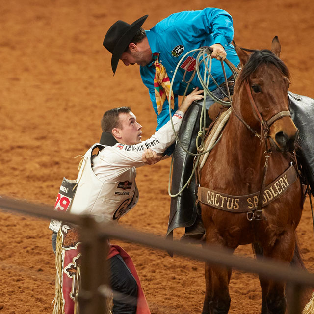 A pickup man leaning over to help cowboy, Tim O’Connell down after his ride.