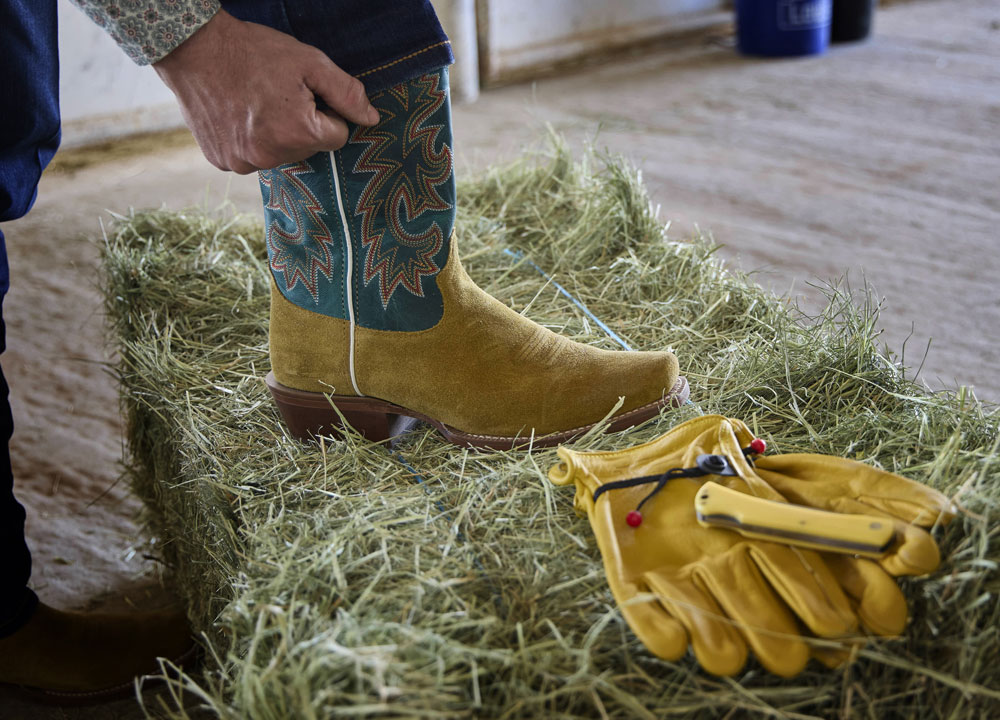 Jimmy 13” Western in wheat suede posed on a hay bale next to a pair of yellow gloves.