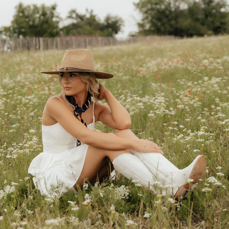 A woman wearing the Justin Evelyn 15-inch Western Boots in Snow (white) while sitting in a field of wildflowers. Shop Evelyn.