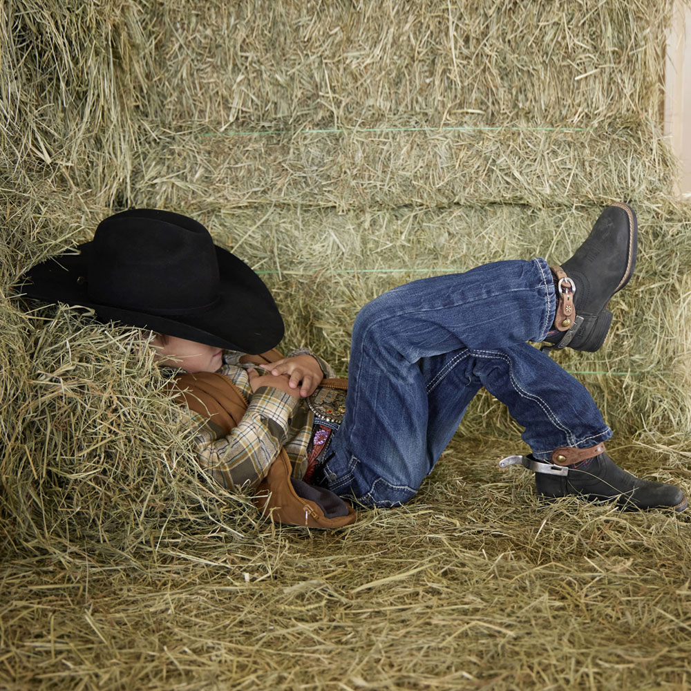 A kid laying down in a hay barn wearing the Bowline Junior boots in black. Shop Bowline Junior