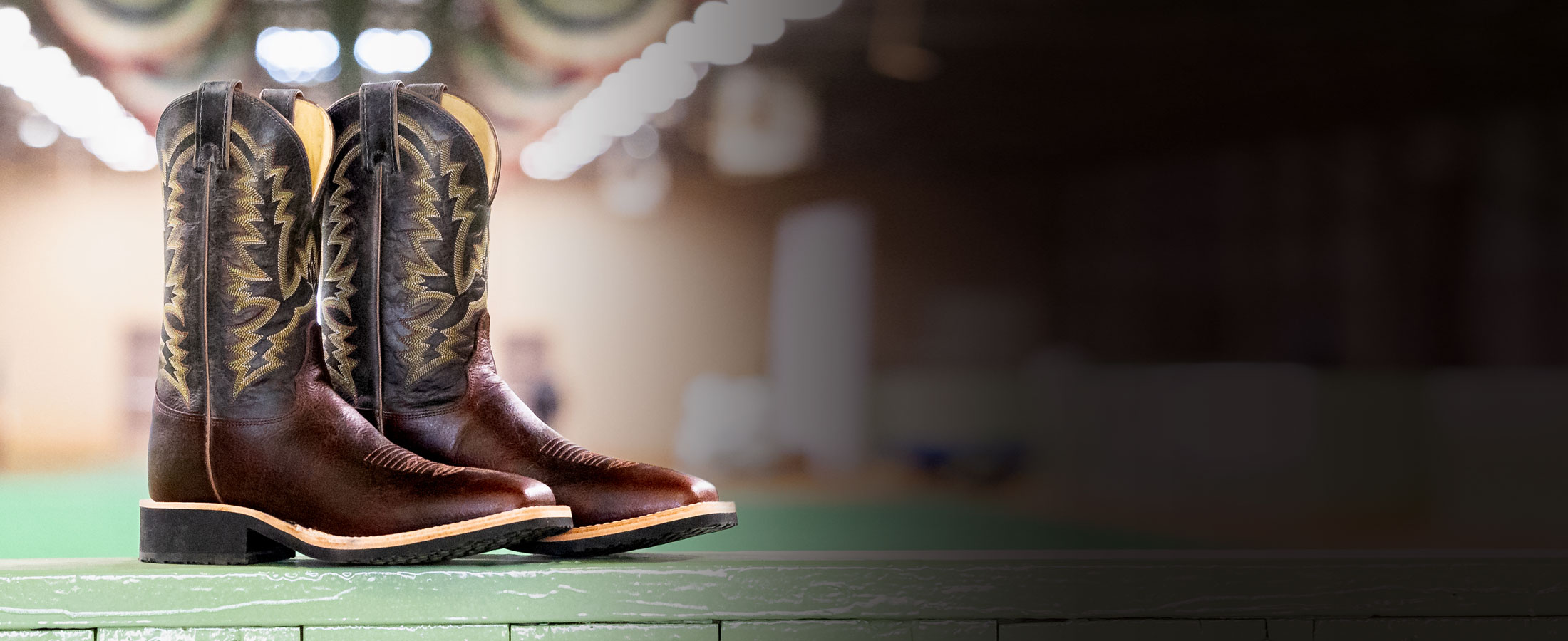 Men’s Trainor 11” Western cowboy boot in brown, posed on a green wood fence with a green show arena and red, white, and blue banners hanging from the ceiling.