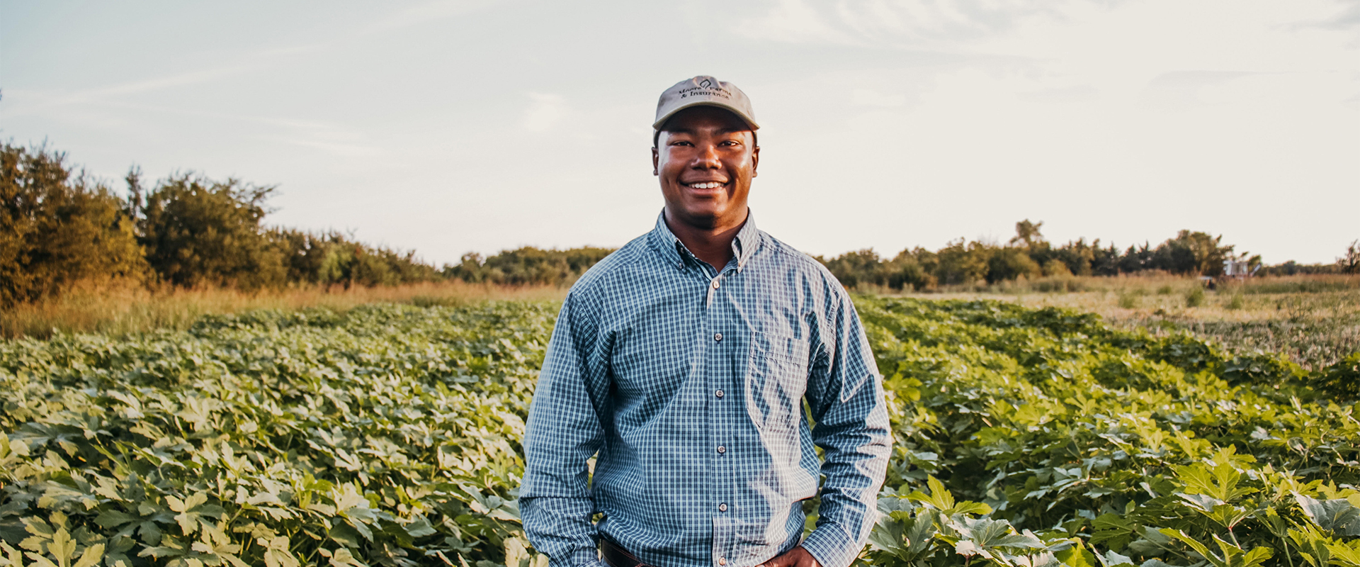 Terren standing in field of crops wearing button up and hat