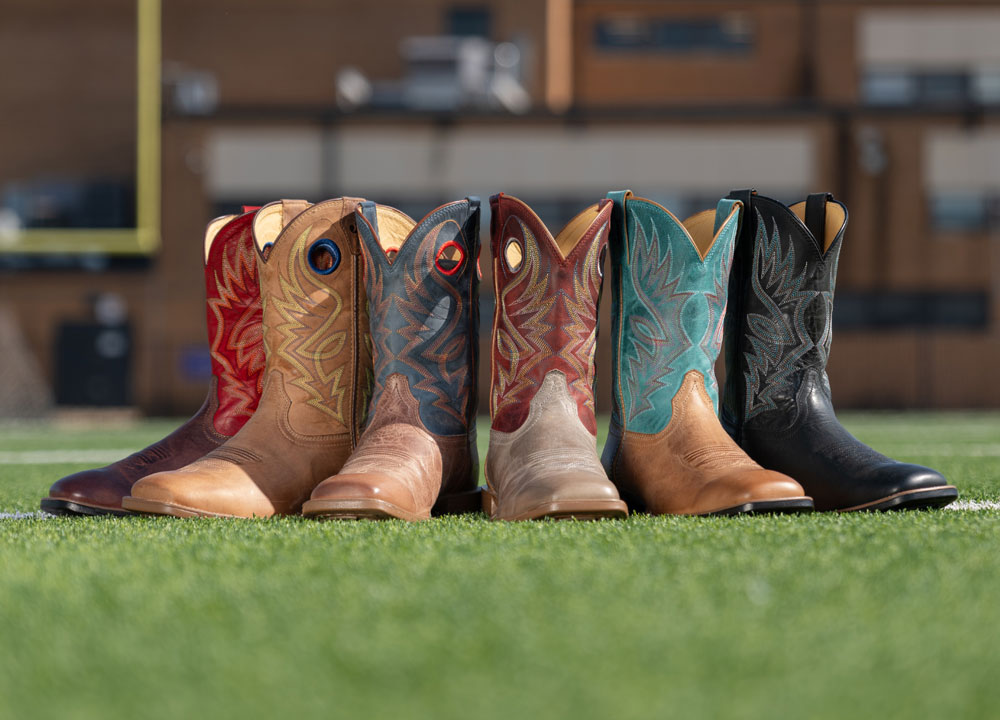 Zeke 11" Western boot in Mohogany, Pickett 11" Western boot in light oak (tan), Pickett 11" Western boot in peanut (brown), Pickett 11" Western boot in Griege (grey), Zeke 11" Western boot in peanut (brown)and Zeke 11" Western boot in peanut (black) on a turf field.