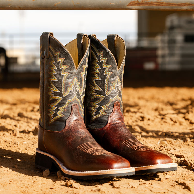 A pair of Justin Trainor 11-inch Western boots in black and dark brown sitting in the sand. Shop Trainor