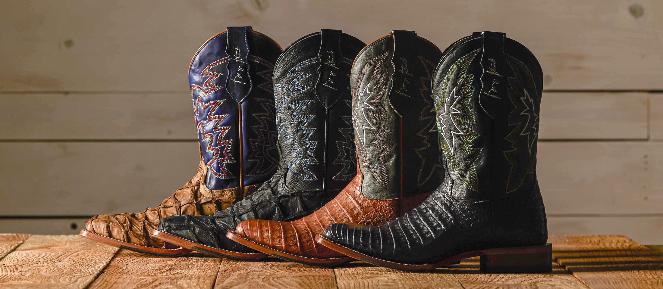 Four boots lined up in a row with their toes to the left and heels to the right with a spotlight on them and placed in front of a wooden backdrop.