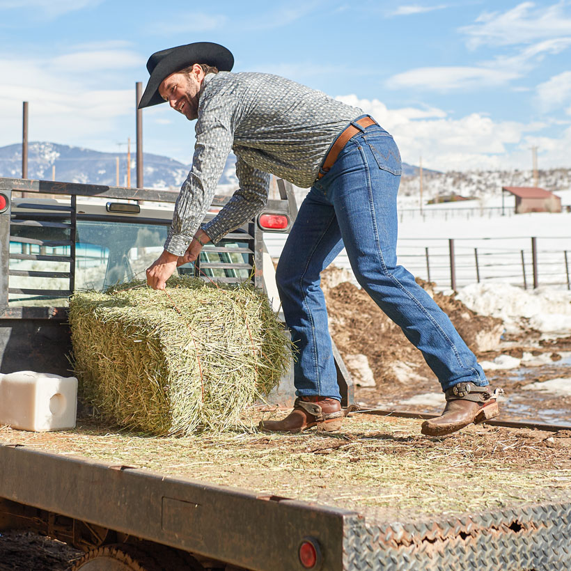 A man standing on the back of a flatbed truck holding a hay bale, smiling.