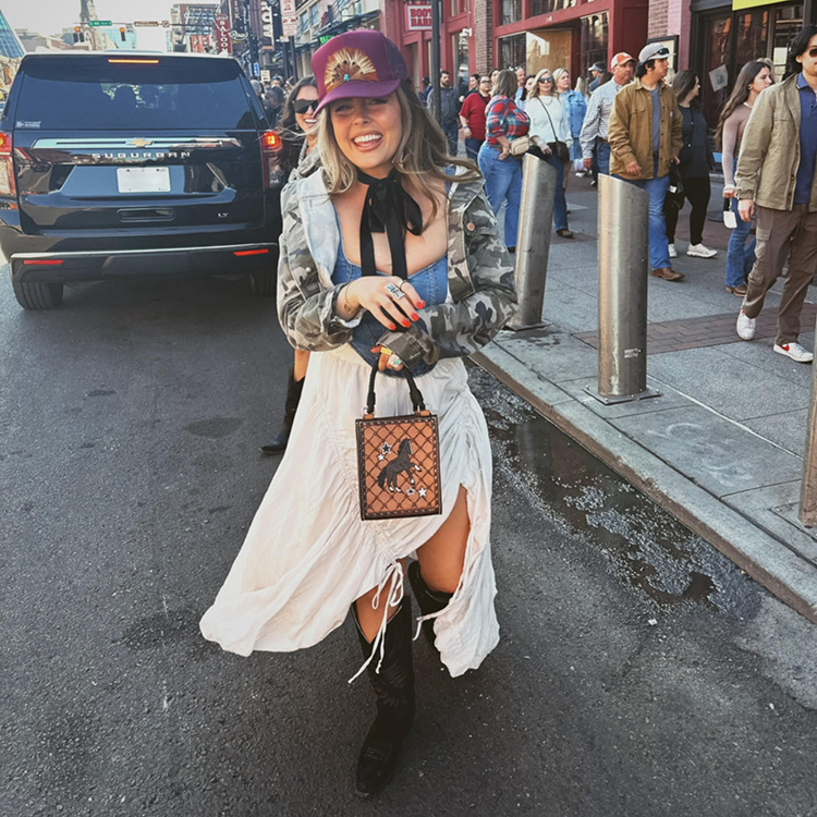 A woman wearing black western boots, a white shirt, and a red baseball cap standing in the street.