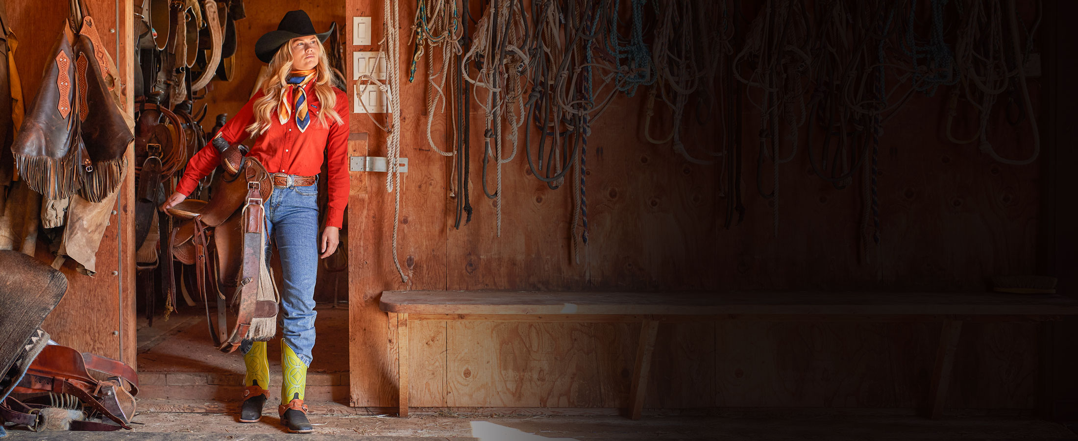 A blonde woman wearing Minick 13” Pull-On Women’s Boot in Lime Green, holding a saddle, inside a barn. 