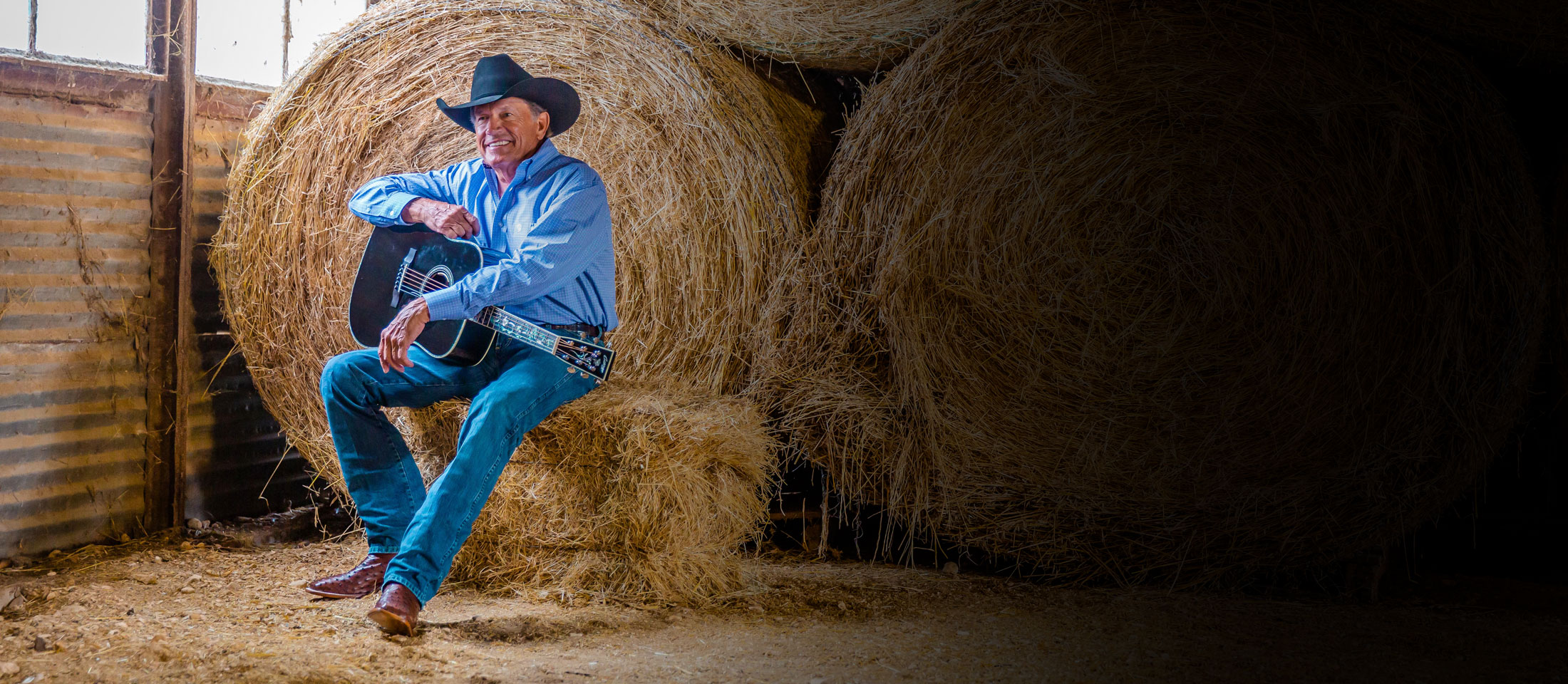 A man wearing Justin George Strait western boots, a cowboy hat and holding a guitar while sitting on a hay bale in a barn.