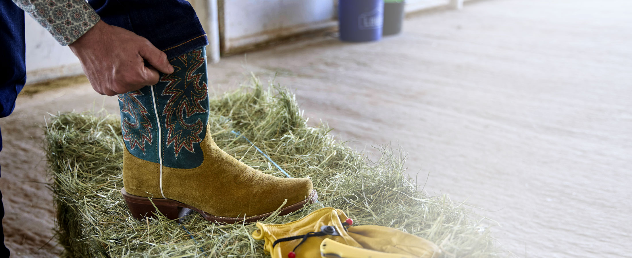 Jimmy 13” Western in wheat suede posed on a hay bale next to a pair of yellow gloves.