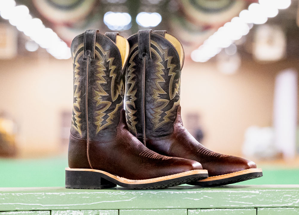 Men’s Trainor 11” Western cowboy boot in brown, posed on a green wood fence with a green show arena and red, white, and blue banners hanging from the ceiling.