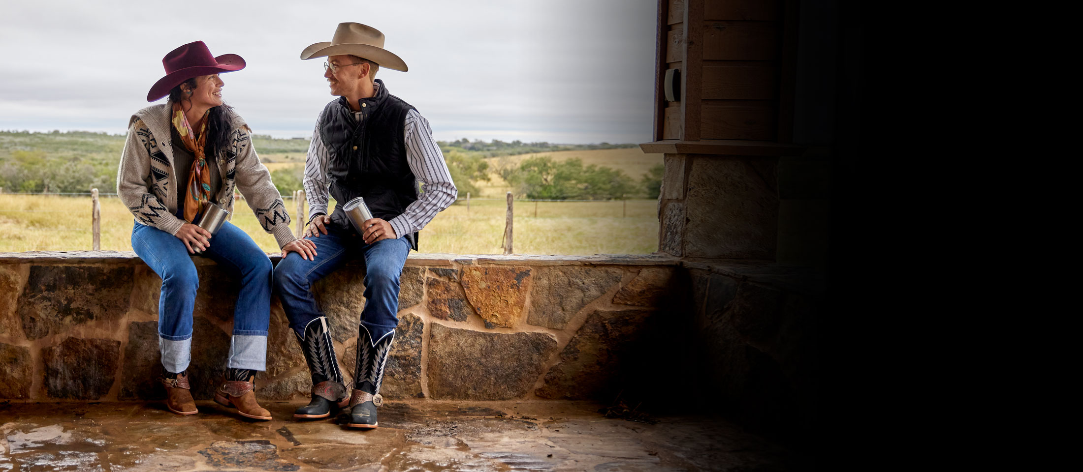 A woman and a man wearing Justin Suede western Boots and cowboy hats while sitting a  wall made of stone.