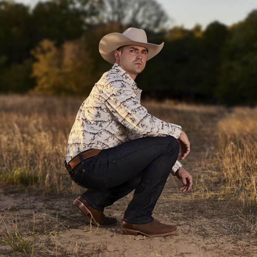 Men’s Classic Straight Leg Jeans in dark blue worn by a man in a cowboy hat kneeling down and posing in a field.