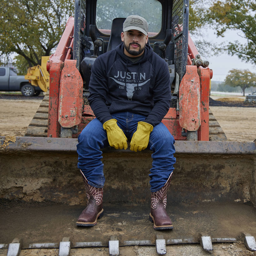 A man wearing Derrickman 12" Waterproof and Performance Fleece Hoodiesitting on a tractor bucket.