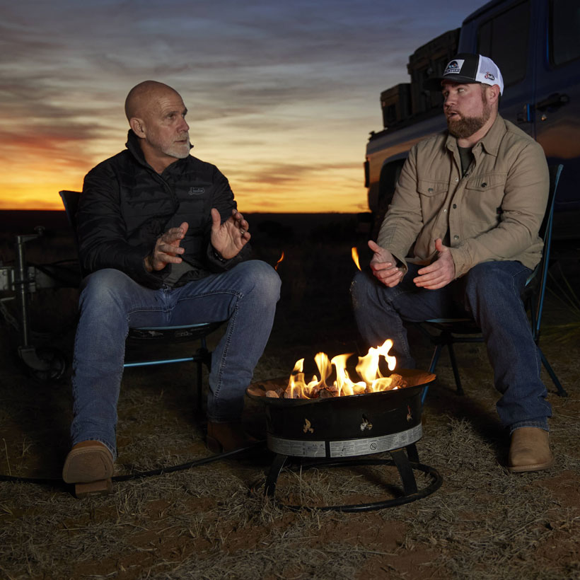 Two men by a fire wearing 1879 Relaxed Bootcut men's Jeans in medium and dark wash.