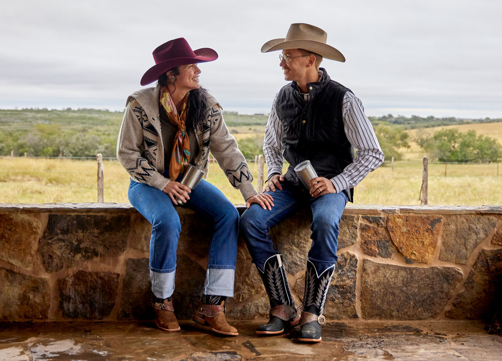 A woman and a man wearing Justin Suede western Boots and cowboy hats while sitting a  wall made of stone.