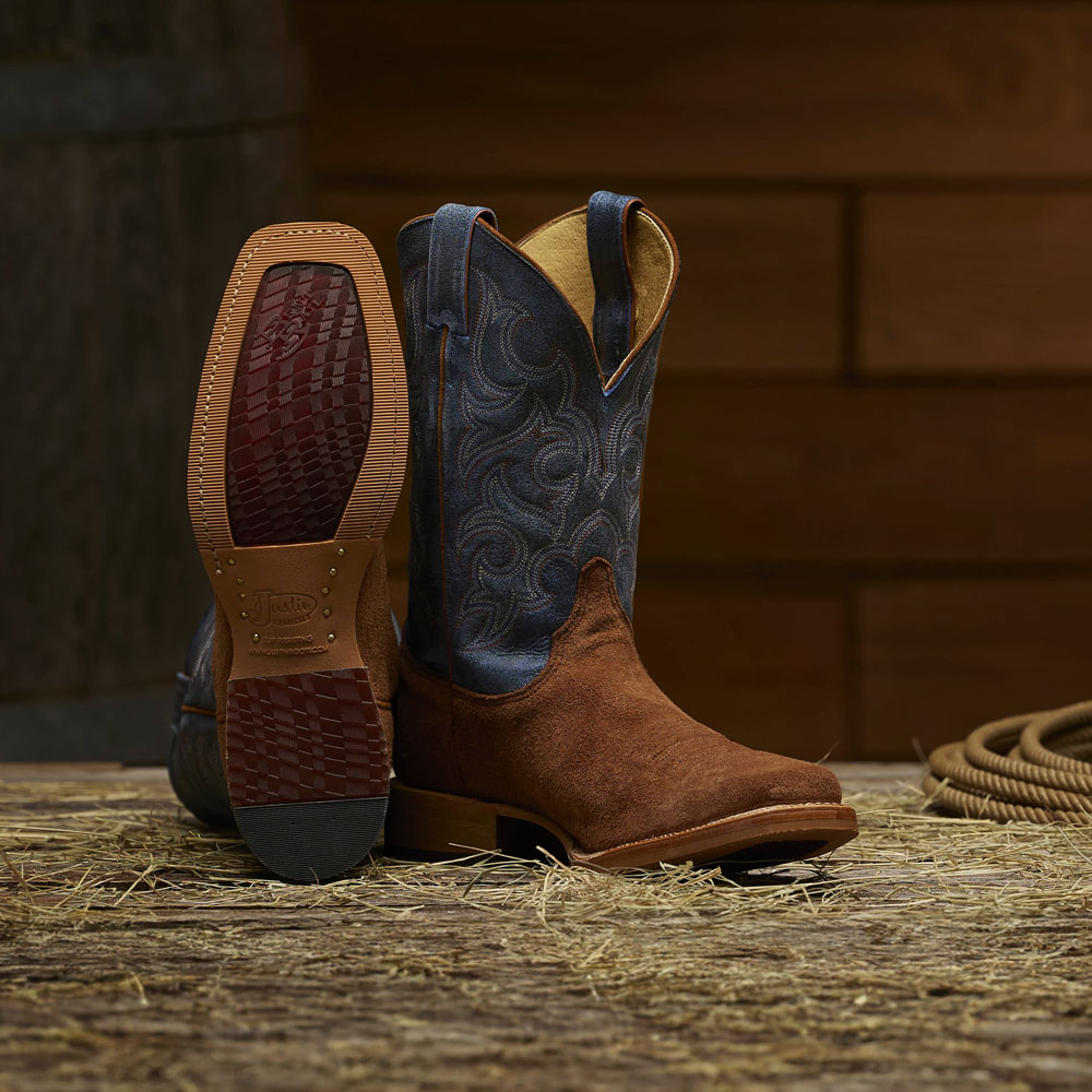 The Justin Fergus western boot in Golden Oak (brown) sitting on a wood floor in a barn. Shop Fergus