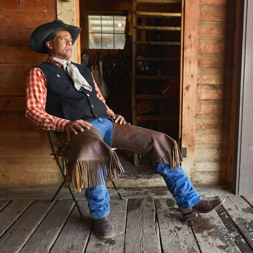 Chaqueta Pascoe de 13” de avestruz suave para hombre en color marrón, usada por un vaquero con sombrero de vaquero y polainas, sentado en una silla.